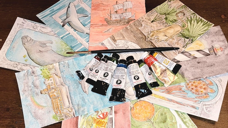 May include: A collection of watercolor paintings featuring marine life, a sailboat, and a lighthouse. A paintbrush and tubes of paint are arranged on top of the paintings. The paintings are in various sizes and colors, with a focus on aquatic themes.