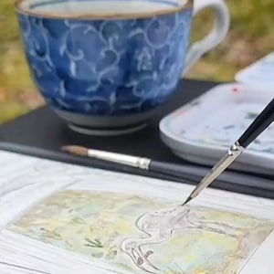 May include: A watercolor painting in progress, featuring a detailed sketch of a bird. A hand holds a fine-tipped paintbrush, applying color to the artwork. A blue and white patterned teacup and a palette of paints are in the background.