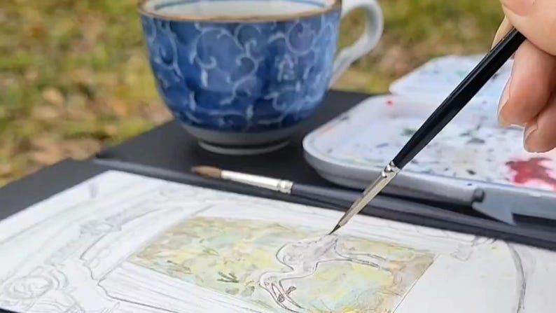 May include: A watercolor painting in progress, featuring a detailed sketch of a bird. A hand holds a fine-tipped paintbrush, applying color to the artwork. A blue and white patterned teacup and a palette of paints are in the background.