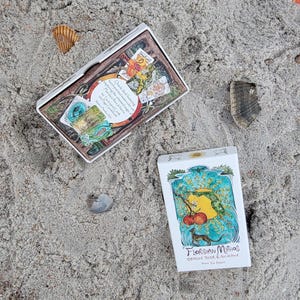 May include: Two white boxes of oracle cards on sand. The top box is open, revealing colorful cards. The bottom box has a watercolor illustration of a yellow shape, red fruit, and a small animal. Text on the box reads "Floridian Mythos Oracle Deck & Guidebook."