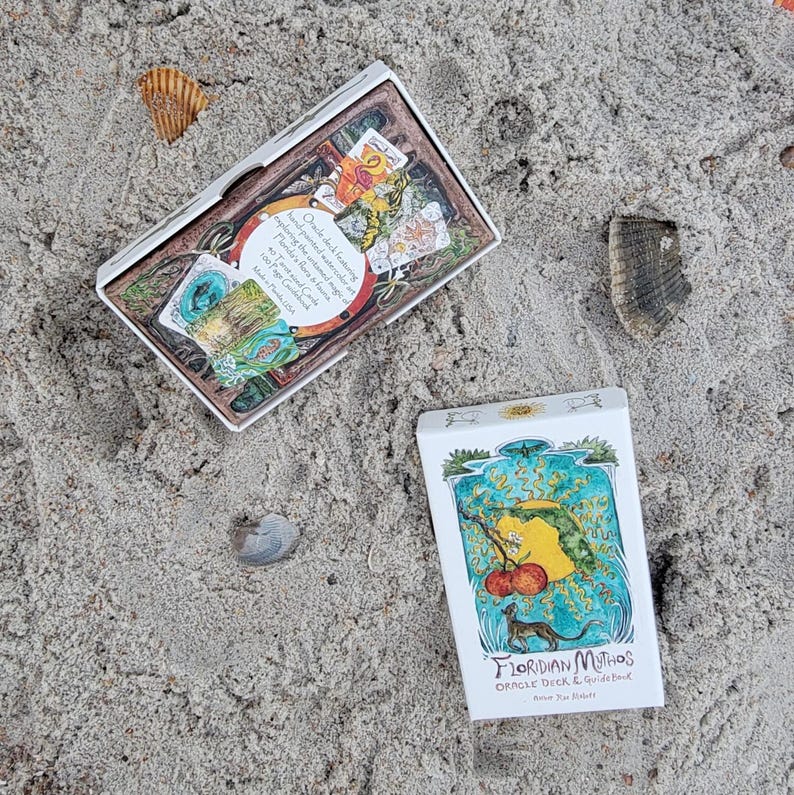 May include: Two white boxes of oracle cards on sand. The top box is open, revealing colorful cards. The bottom box has a watercolor illustration of a yellow shape, red fruit, and a small animal. Text on the box reads "Floridian Mythos Oracle Deck & Guidebook."