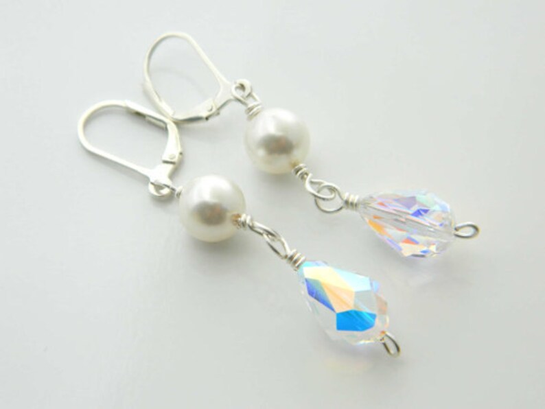 May include: Dangle earrings with a white pearl and a clear, iridescent crystal teardrop. The earrings have silver-coloured lever-back closures and are connected with silver wire.