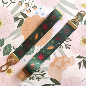 May include: Two dark green lanyards with gold-colored hardware and a fall-themed pattern of leaves, acorns, and berries. The lanyards are lying on a floral patterned background. The lanyards are approximately 1 inch wide.