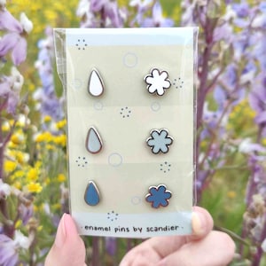 May include: A set of six enamel pins in a clear plastic package. The pins feature raindrop and flower designs in white, light blue, and dark blue with silver-colored metal borders. The text "enamel pins by scandier" is printed on the package.