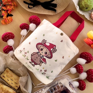 Könnte beinhalten: Eine cremefarbene Tragetasche mit roten Griffen, verziert mit einer Cartoonfigur mit einem Pilzhut. Das Bild enthält pilzförmige Dekorationen, Blumen und Gebäck. Auf der Tasche steht der Text "scandier".