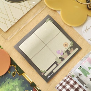 May include: A square notepad with a dark gray border and a light beige interior divided into four sections. The top right corner features a small illustration of potted flowers. The bottom left corner has the word "DATE:" printed on it. Other stationery items are in the background.