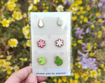 Puede incluir: Un conjunto de seis pines de esmalte en un paquete transparente. Los pines presentan una lágrima blanca, una lágrima verde claro, dos flores rosas, una hoja de nenúfar verde y una rana verde. El texto "enamel pins by scandier" está en la parte inferior.