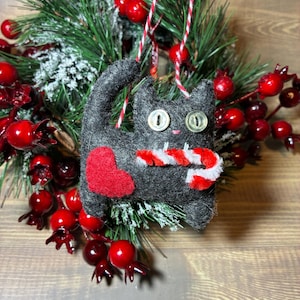 May include: A handmade felt cat Christmas ornament. The gray cat has button eyes, a red heart, and a candy cane scarf. It hangs from a red and white twine string. The ornament is surrounded by a wreath of green pine needles and red berries.