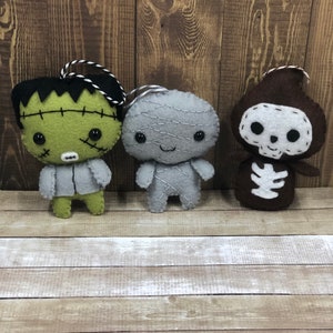 May include: Three felt Halloween ornaments, a green Frankenstein, a gray mummy, and a brown skeleton. Each ornament has a stitched-on face and a loop for hanging.