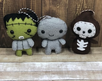 May include: Three felt Halloween ornaments, a green Frankenstein, a gray mummy, and a brown skeleton. Each ornament has a stitched-on face and a loop for hanging.