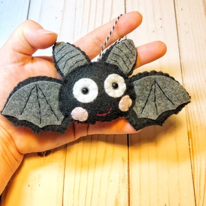May include: A gray felt bat ornament with black eyes, a pink nose, and a red stitched mouth. The bat has a black and white string attached to the top for hanging.