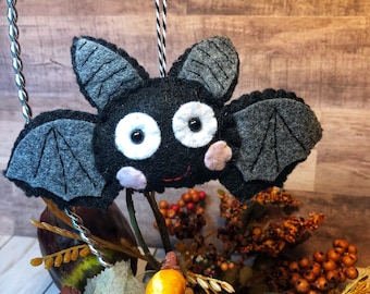 May include: A black felt bat with gray wings and white eyes, hanging from a string. The bat has a smiling mouth and pink cheeks. The bat is hanging in front of a brown wooden background.