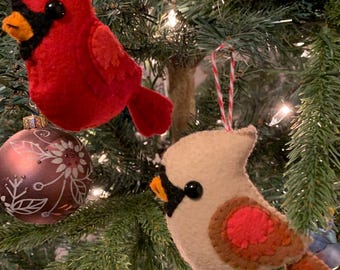 May include: Two felt cardinal ornaments hanging on a Christmas tree. One cardinal is red with a black eye and the other is beige with a red breast and black eye.