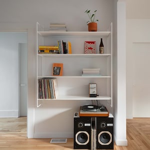 May include: A white bookshelf with four shelves, filled with books, records, and a small radio. A turntable sits on top of a set of black speakers.