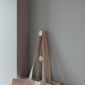 Peut inclure: Un sac fourre-tout beige avec du texte noir suspendu à deux crochets blancs sur un mur gris clair. Le texte sur le sac est "FRGMNT, Jimmy Gonzalez, Micha Martini, Mara Dupas, Joliz Dela Peña, Thea Patterson et Rachel Harris et Elinor Fueter, Mycelium, Louise Michel Jackson, Vat..."