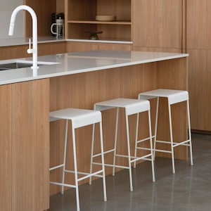 May include: Three white metal bar stools with a simple, modern design. The stools are arranged in a row in front of a kitchen island with a white worktop and wood cabinets.