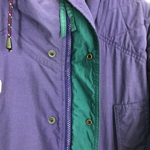 May include: Close-up of a purple and green jacket. The jacket features a zipper, snap closures, and a drawstring hood. The jacket has a quilted design and a pocket. The color scheme includes purple, green, and a hint of white.