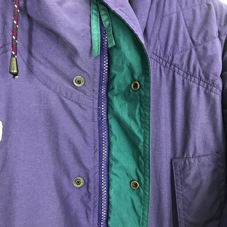 May include: Close-up of a purple and green jacket. The jacket features a zipper, snap closures, and a drawstring hood. The jacket has a quilted design and a pocket. The color scheme includes purple, green, and a hint of white.