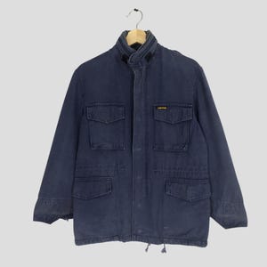 May include: A dark blue utility jacket with a high collar, multiple pockets, and a drawstring waist. The jacket has a small yellow label with the text "Carhartt" on the left chest.