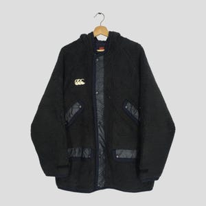 May include: Black hooded jacket with a fuzzy texture, featuring a quilted panel down the center and on the pockets. The jacket has a gold logo on the chest and is buttoned up. The jacket is hanging on a wooden hanger.