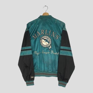 May include: Teal and black satin jacket featuring the Miami Marlins logo and text. The jacket has black sleeves with teal stripes and the words "Major League Baseball" below the logo. The jacket is hanging on a wooden hanger.