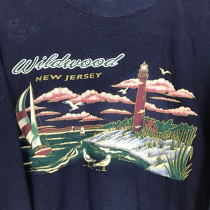Może przedstawiać: Granatowa bluza z nadrukiem graficznym latarni morskiej, żagl&oacute;wki i sceny plażowej. Na bluzie nadrukowany jest tekst "Wildwood New Jersey".