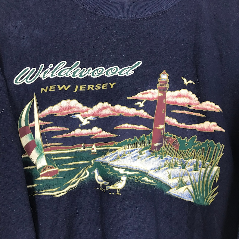 Może przedstawiać: Granatowa bluza z nadrukiem graficznym latarni morskiej, żagl&oacute;wki i sceny plażowej. Na bluzie nadrukowany jest tekst "Wildwood New Jersey".