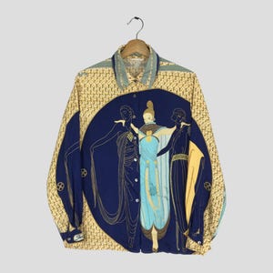 May include: A button-down shirt with a unique design. The shirt features a yellow background with a repeating pattern, blue sleeves, and a large blue circle on the front. Inside the circle are three figures in flowing gowns. The shirt is hanging on a wooden hanger.