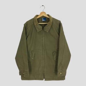 Peut inclure: Veste vert olive avec un col en velours côtelé marron, une fermeture éclair sur toute la longueur et deux poches avant. La veste a des poignets boutonnés et est suspendue à un cintre en bois.