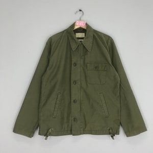 May include: Olive green jacket with a button-up front, a collared neckline, and a single chest pocket. The jacket has long sleeves and adjustable drawstrings at the hem. The jacket is hanging on a metal hanger.