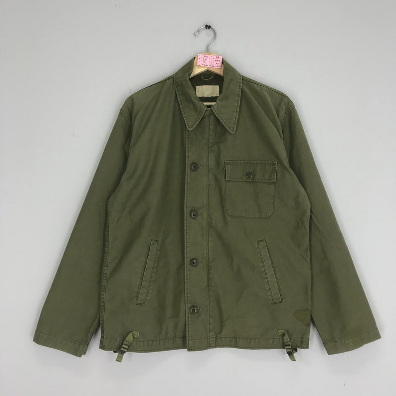 May include: Olive green jacket with a button-up front, a collared neckline, and a single chest pocket. The jacket has long sleeves and adjustable drawstrings at the hem. The jacket is hanging on a metal hanger.
