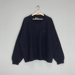 Könnte beinhalten: Ein marineblauer Pullover mit einem kleinen gestickten Logo auf der linken Brust. Das Logo ist ein weißer Stern in einem schwarzen Kreis.