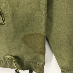 May include: Close-up of an olive green jacket. The fabric has a textured appearance. A drawstring and a small pocket are visible. There is a stain on the jacket. The jacket's sleeve is also visible.
