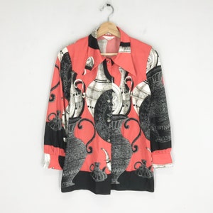 Puede incluir: Una camisa de manga larga de color rosa coral con botones y un estampado en blanco y negro de jarrones estilizados. La camisa tiene un cuello ancho y una banda negra en la parte inferior.