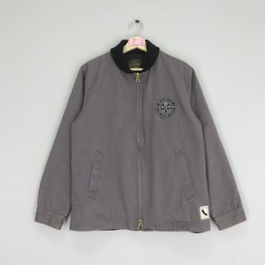 Puede incluir: Una chaqueta de trabajo gris con una cremallera negra y un cuello negro. La chaqueta tiene un gran parche circular en la parte delantera con el texto "WORKERS OF THE WORLD UNITE" y un parche más pequeño en la manga con el texto "CALIFORNIA"