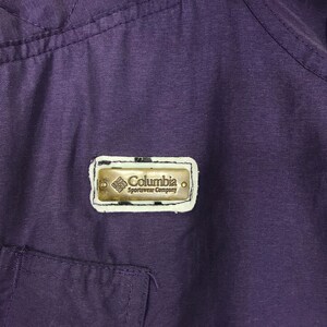 May include: Close-up of a deep purple garment with a small pocket and stitching details. A rectangular metal Columbia Sportswear Company logo is affixed to the fabric, featuring the brand name and logo.