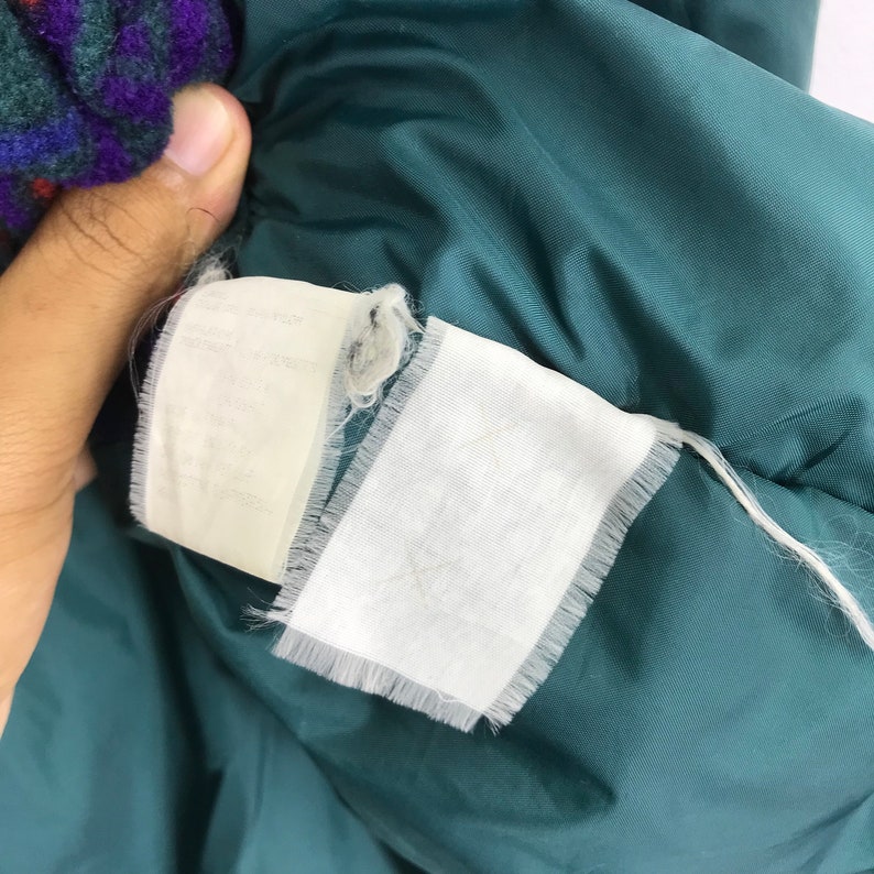 May include: Close-up of a teal-colored jacket interior with two white fabric tags. The tags have frayed edges and appear to have text. A portion of a patterned fabric is visible in the upper left corner.