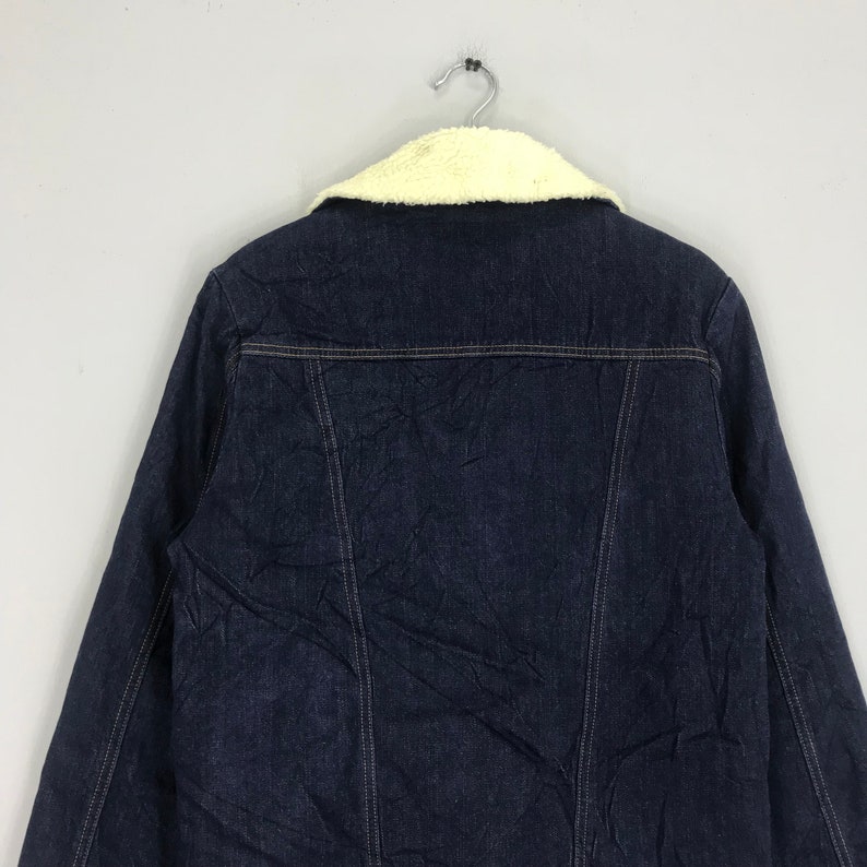 Op de afbeelding: Een donkerblauw denim jack met een witte nep bontkraag. Het jack heeft een knoopsluiting en twee zakken aan de voorkant.