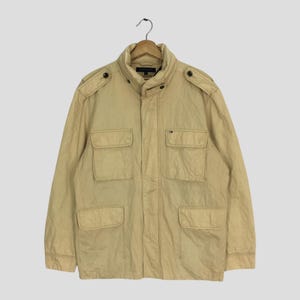 May include: A tan jacket with a high collar and four front pockets. The jacket has shoulder epaulets and a small logo on the chest. It is hanging on a wooden hanger.
