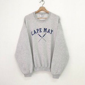 Puede incluir: Una sudadera gris con un gráfico de remos cruzados en azul marino y rojo y el texto "CAPE MAY" en azul marino.