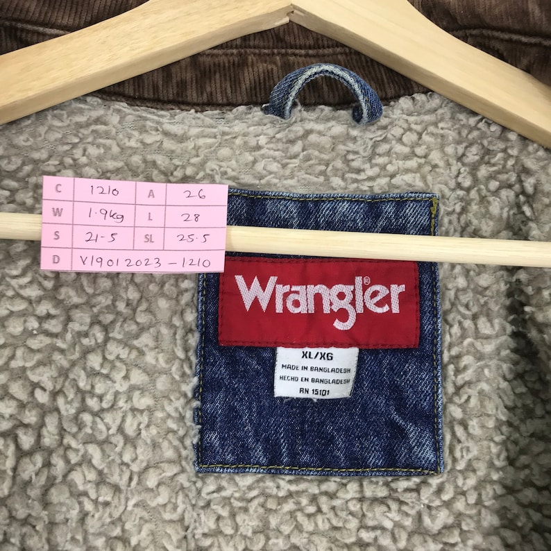 Puede incluir: Una chaqueta vaquera azul con cuello de pana marr&oacute;n y forro polar. La chaqueta tiene una etiqueta de Wrangler con la talla XL/XG y el texto "MADE IN BANGLADESH" y "RN 15101".
