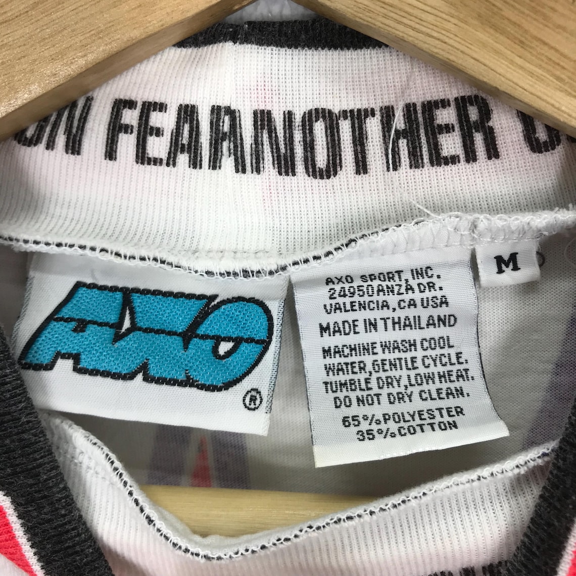 May include: White t-shirt with a blue and black logo that says "AVO" and the text "IN FEARNOTHER" on the collar. The tag says "AXO SPORT, INC. 24950 ANZADR. VALENCIA, CA USA MADE IN THAILAND MACHINE WASH COOL WATER, GENTLE CYCLE. TUMBLE DRY, LOW HEAT. DO NOT DRY CLEAN. 65% POLYESTER 35%COTTON" and has a size "M" tag.