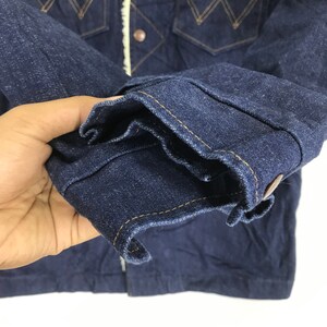 Op de afbeelding: Close-up van de donkerblauwe denim manchet van een jas met een enkele knoop en gele stiksels.