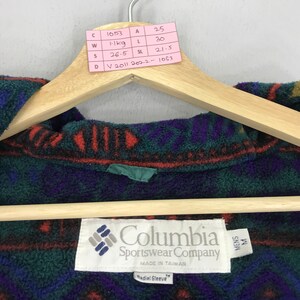 May include: A close-up of a Columbia Sportswear Company fleece jacket. The jacket features a geometric pattern in green, purple, and red. The tag reads "Mens M" and "Made in Taiwan". The jacket is hanging on a wooden hanger.