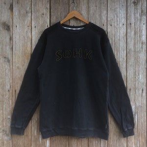 Könnte beinhalten: Schwarzer Pullover mit einem gelben gestickten "SOHK"-Logo auf der Vorderseite.