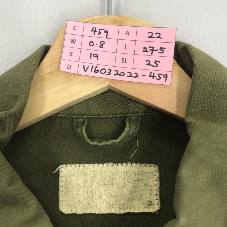 May include: Olive green jacket hanging on a wooden hanger. The jacket has a collar and a small fabric label. A pink tag with black text is pinned to the hanger, displaying measurements and a product code.