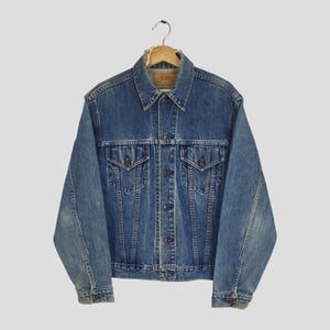 Puede incluir: Chaqueta vaquera azul vintage con un diseño clásico. La chaqueta tiene una parte delantera abotonada, dos bolsillos en el pecho y dos bolsillos laterales. La chaqueta está colgada de una percha de madera.