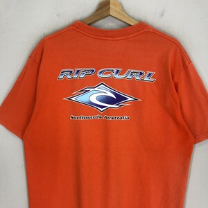 Pu&ograve; includere: T-shirt arancione con un logo Rip Curl blu e bianco e il testo "Surfboards Australia".