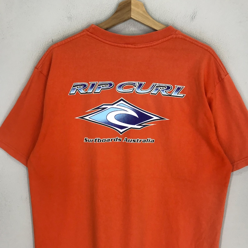 Pu&ograve; includere: T-shirt arancione con un logo Rip Curl blu e bianco e il testo "Surfboards Australia".