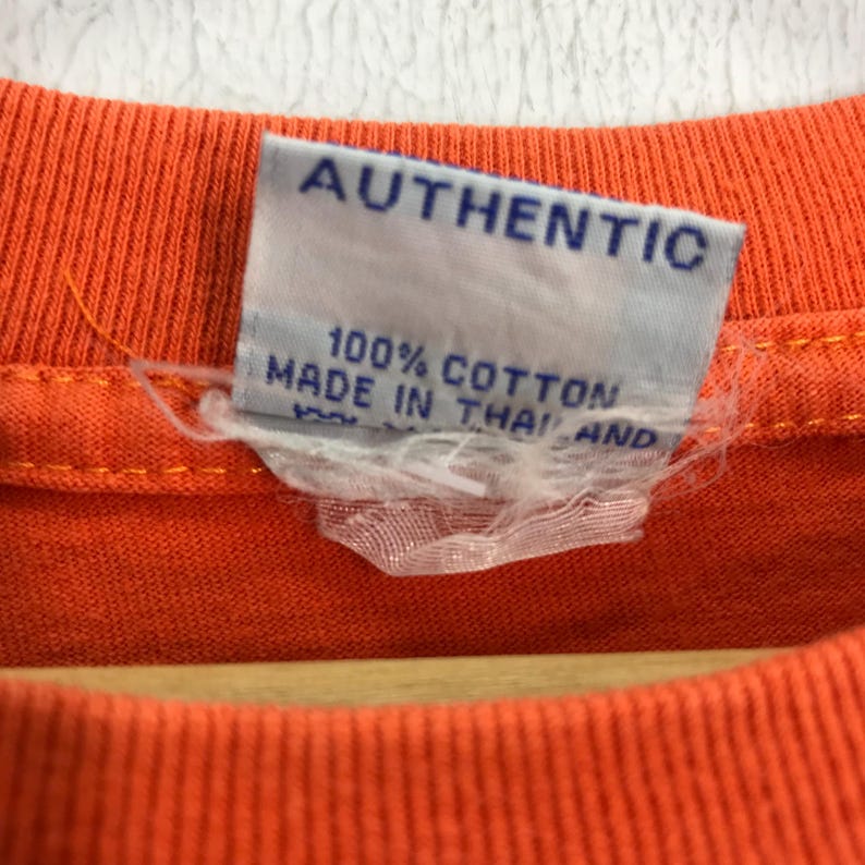 Pu&ograve; includere: Primo piano di una maglietta arancione brillante con un'etichetta bianca che recita "AUTHENTIC 100% COTTON MADE IN THAILAND".